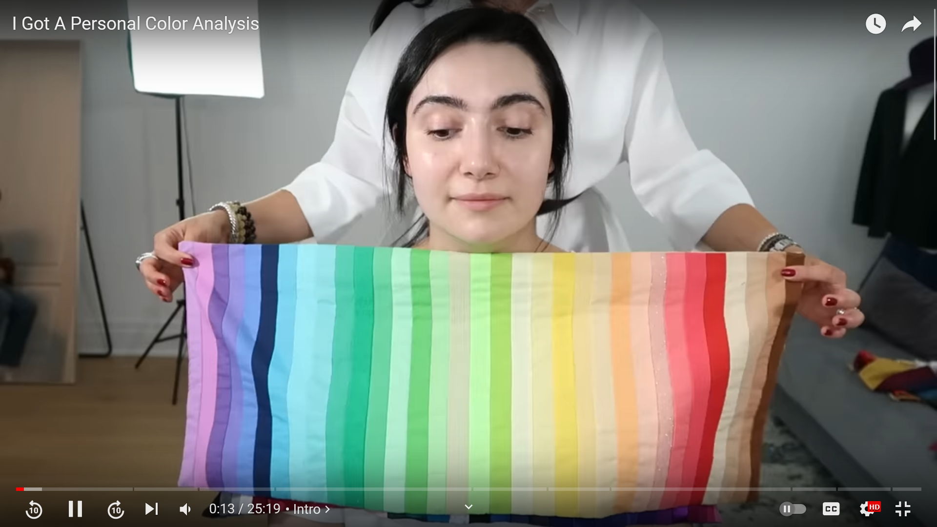 A screenshot of a YouTube video by Safiya Nygaard. She looks down and to the left as somebody behind her holds a piece of cloth with lots of different coloured stripes below her face. Their face is not visible.